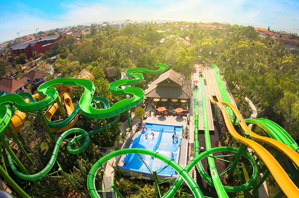 Aerial view of a water park featuring winding green slides, a flow rider surfing attraction, and surrounding greenery, Bali, Indonesia.