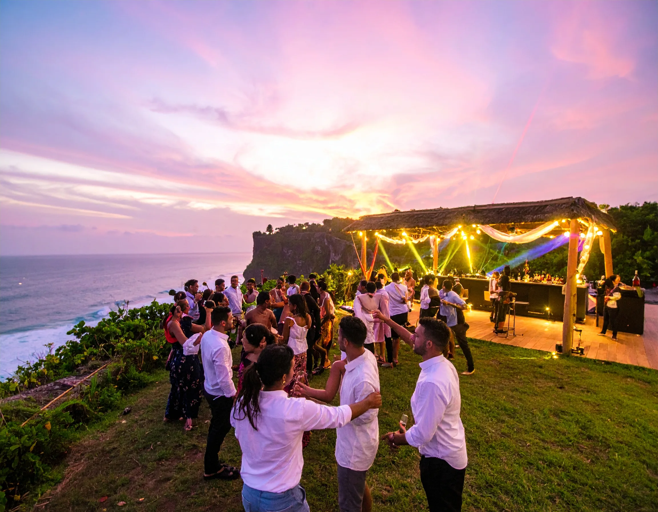 Luxury cliffside club overlooking ocean, champagne bottles, EDM DJ with laser lights, couples dancing, sunset sky turning neon